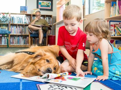 Young boy and girl reading a book to a golden retriever dog who watches them read