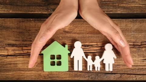 Two hands over a table framing paper cutouts of a family standing next to a house.