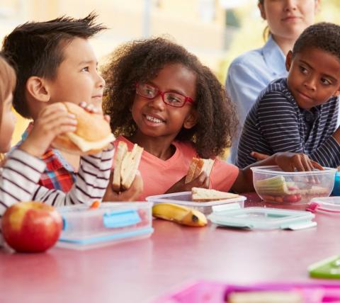 children of various ethnicities eating sandwiches and fruit at a table