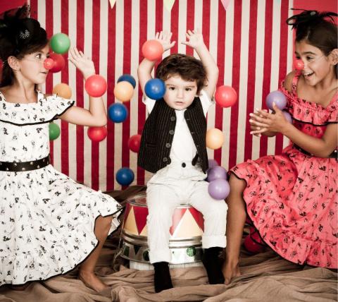 a small boy sitting on a chair juggling balls with a girl on either side of him in front of a striped backghround