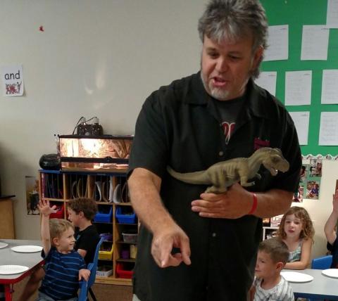 Man holding a dinosaur talking with young children in a classroom