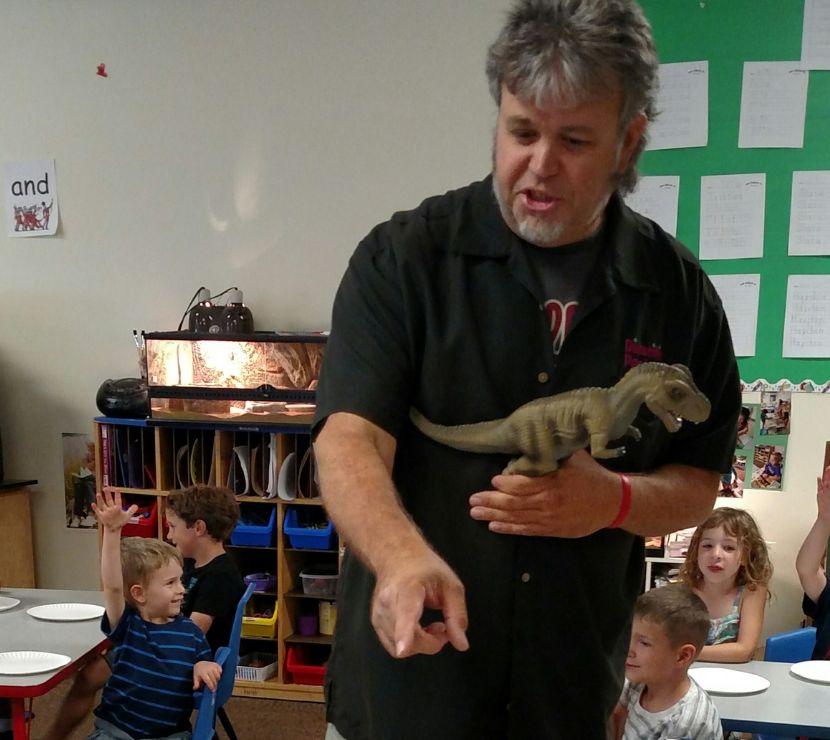 Man holding a dinosaur talking with young children in a classroom